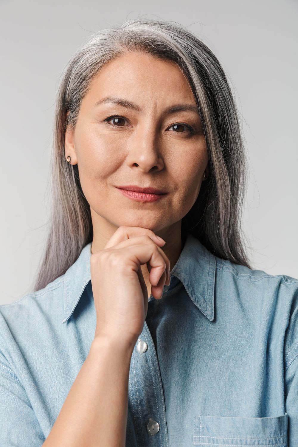 A woman with long gray hair, possibly considering hormone replacement therapy, wears a light blue button-up shirt and looks at the camera with a neutral expression, her hand thoughtfully resting under her chin.